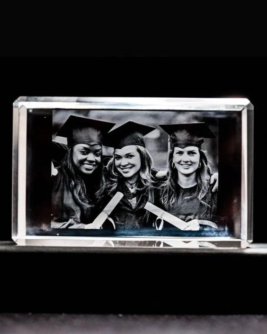 3 graduates inside a glass block, wearing caps and gowns, smiling.